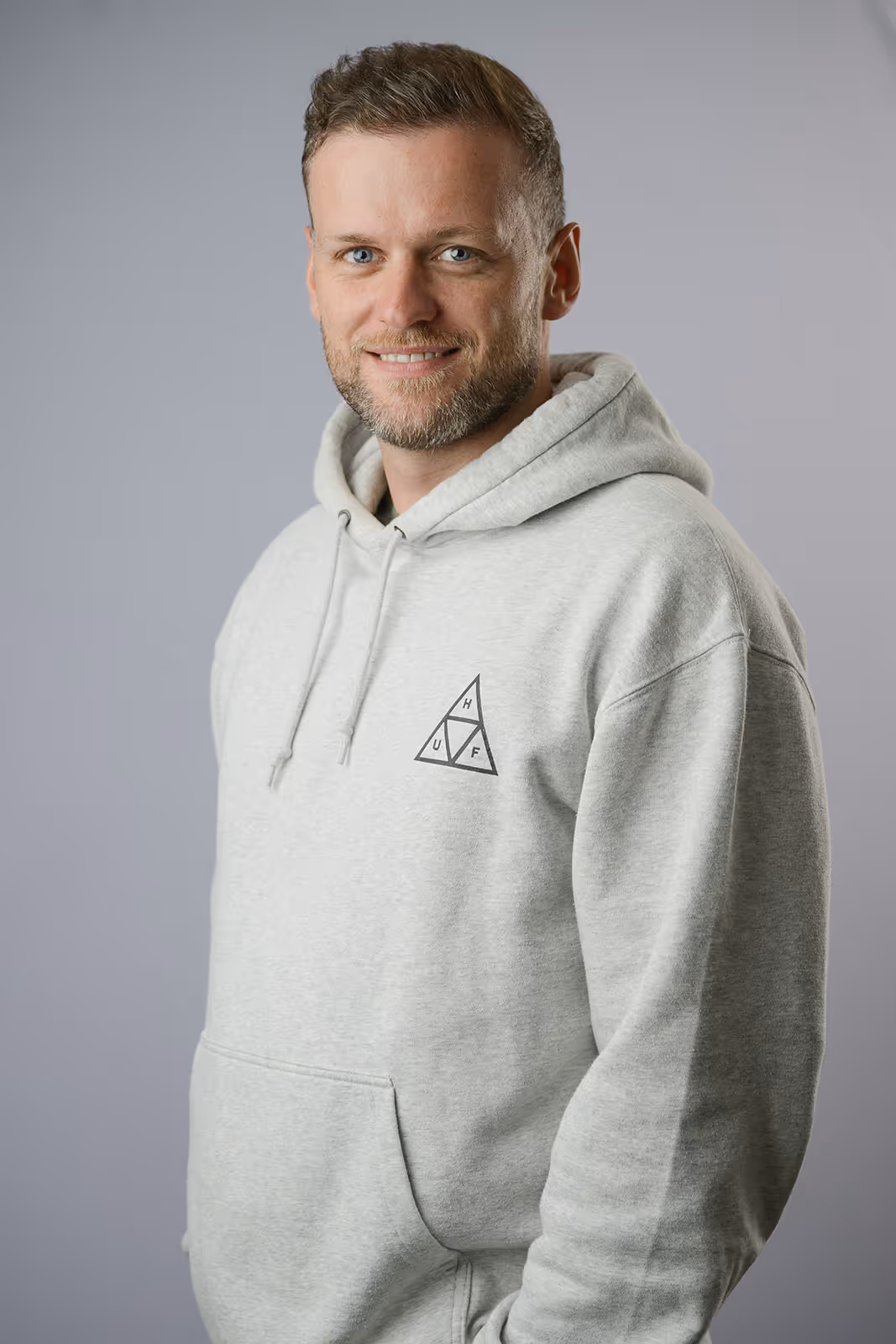 Man with short styled hair and a trimmed beard, wearing a sage green t-shirt, holding a white mug labeled "Harrier" with a playful expression, standing against a light grey background.