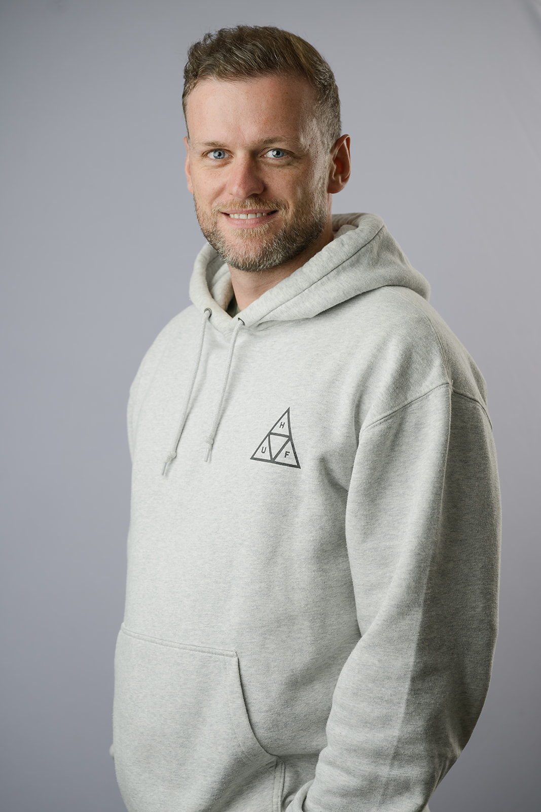 Man with short styled hair and a trimmed beard, wearing a sage green t-shirt, holding a white mug labeled "Harrier" with a playful expression, standing against a light grey background.