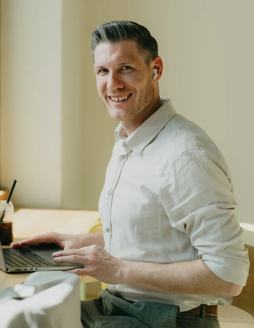 Lächelnder Mann mit weißen Kopfhörern, der an einem Laptop an einem hellen Tisch arbeitet.
