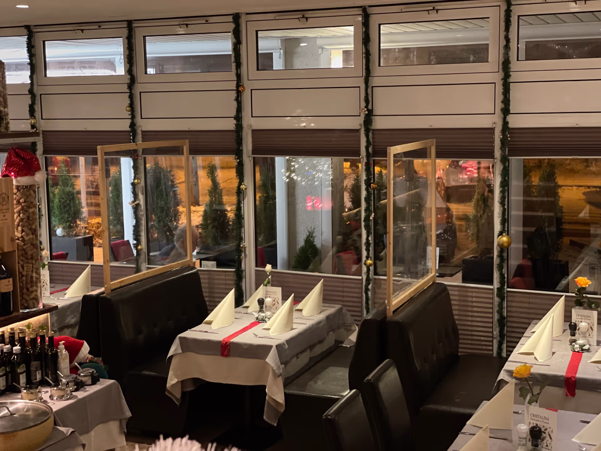 Cozy restaurant interior with tables set with folded napkins, a red table runner, flowers, and clear plastic dividers between seating booths.