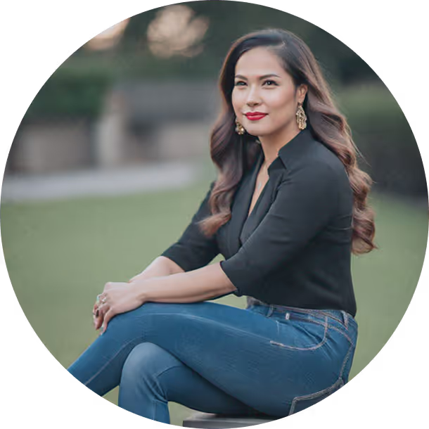 Confident woman sitting outdoors on a stool, wearing a black blouse and blue jeans, softly smiling with arms crossed over her knees.