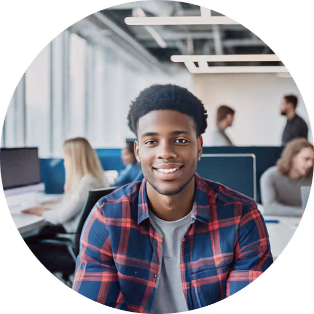 Young man in a plaid shirt smiling at his desk in a bright open-plan office.