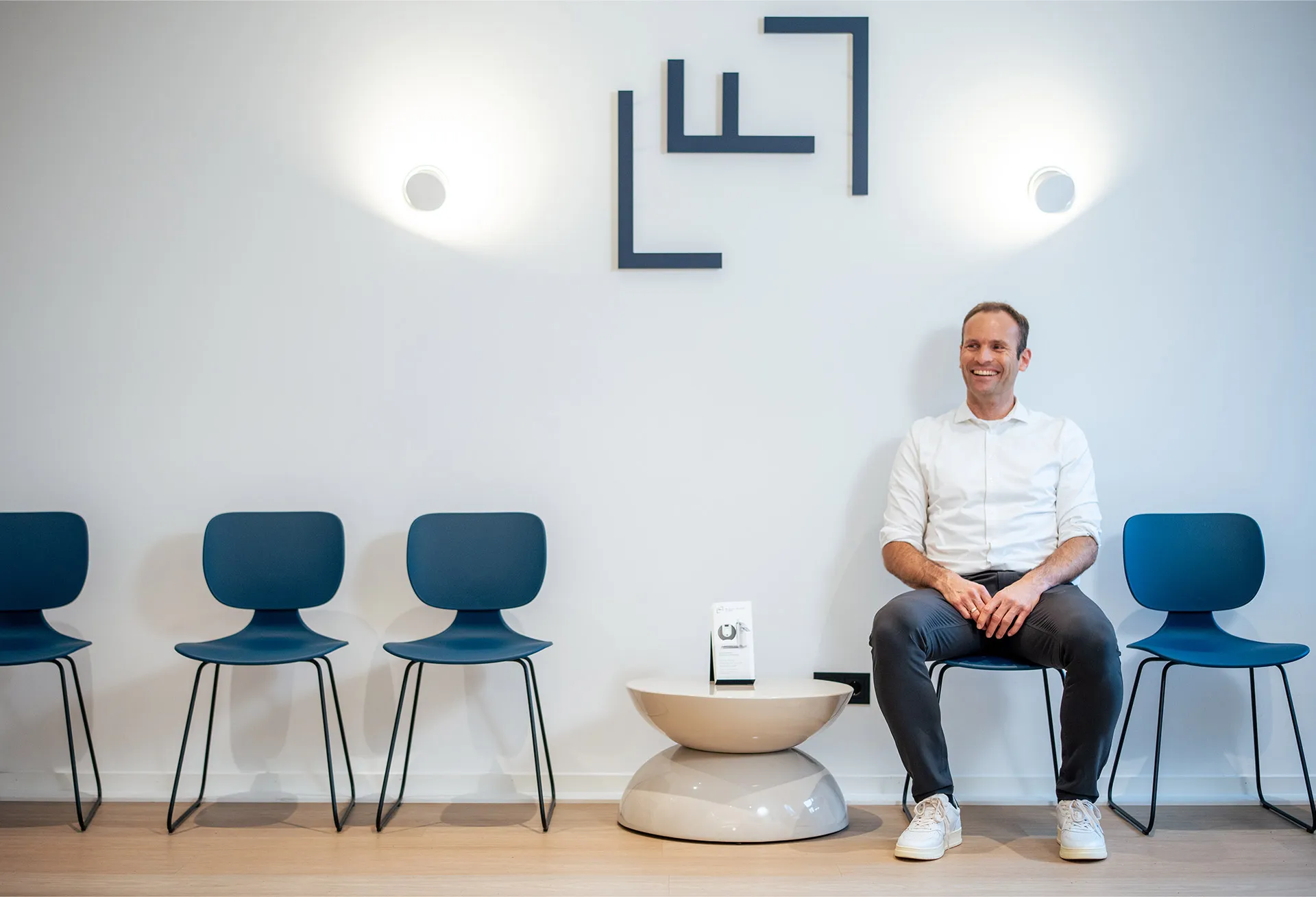 Dr. Fluck sitzt im weißem Hemd, lächelnd im modernen Wartezimmer der Praxis, mit blauen Stühlen und kleinem runden Beistelltisch.