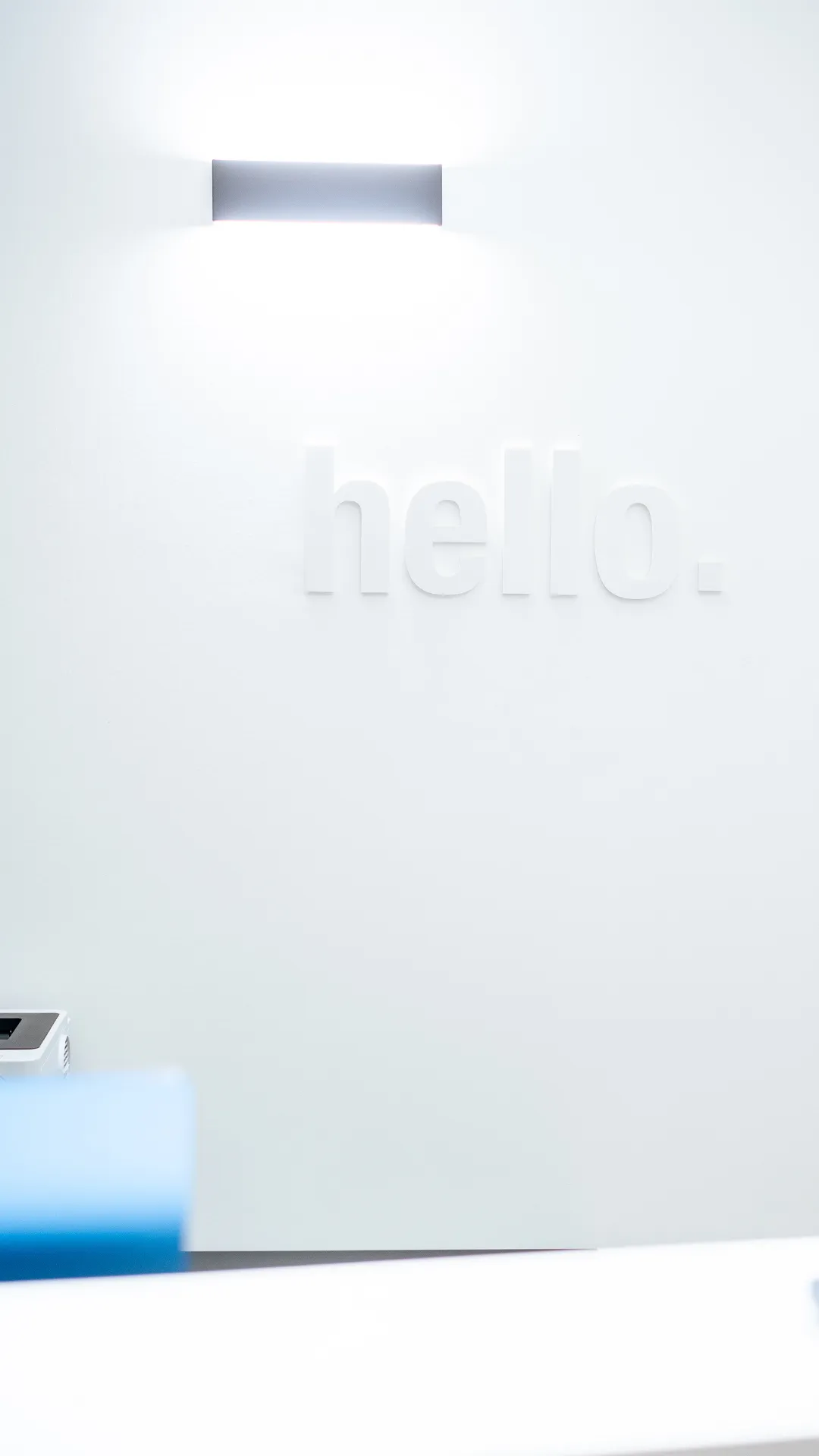 Weiße Wand mit erhabenen Buchstaben, die das Wort ‚hello.‘ zeigen, beleuchtet von einer modernen Wandlampe.