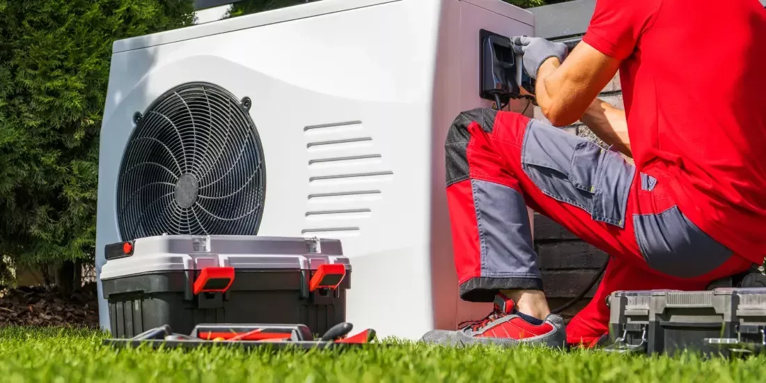 . 10. A technician in a red shirt and grey pants is kneeling and working on the internal components of a white outdoor heat pump unit