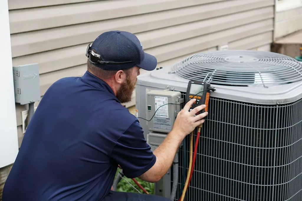 A close-up of a bearded HVAC technician in a dark blue polo and cap, using a digital manifold gauge to check the refrigerant levels of an outdoor heat pump unit.