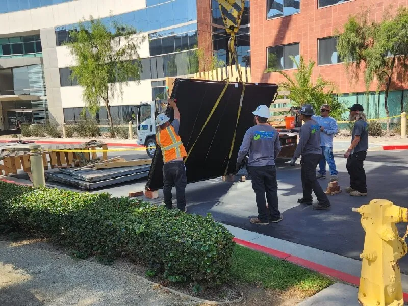 Construction workers using crane to move large black panel near office buildings