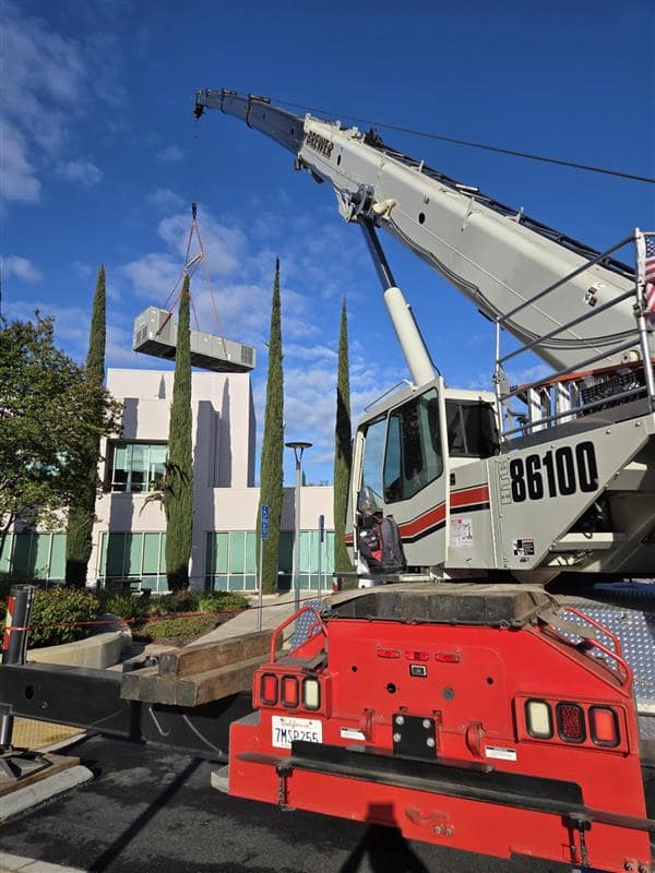 Large mobile crane parked near modern building with cypress trees