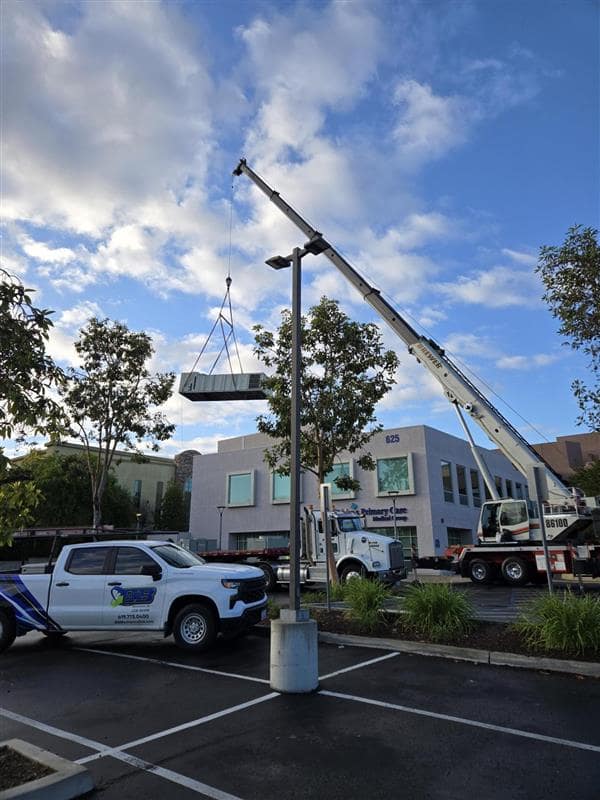 Crane lifting large HVAC unit in parking lot near modern building