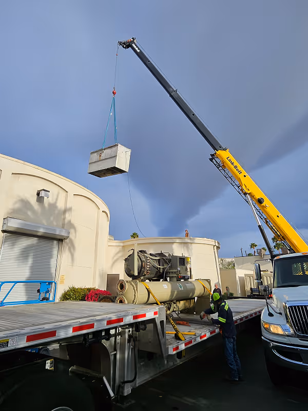 Yellow crane lifting heavy equipment onto flatbed truck near industrial building