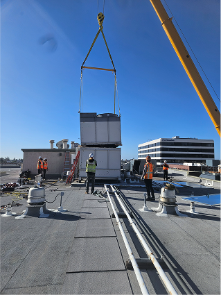 Crane lifting large equipment on rooftop with construction workers