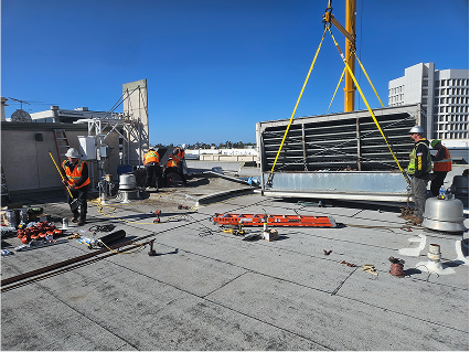 Construction workers using crane to lift large HVAC equipment on rooftop