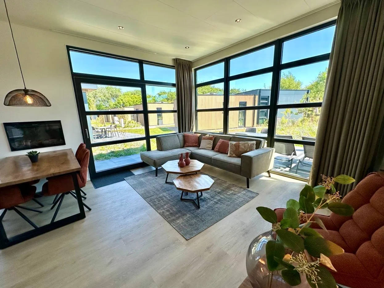 Eckfenster, L-förmiges graues Sofa, Couchtisch, Esstisch, große Fenster und Blick ins Grüne.