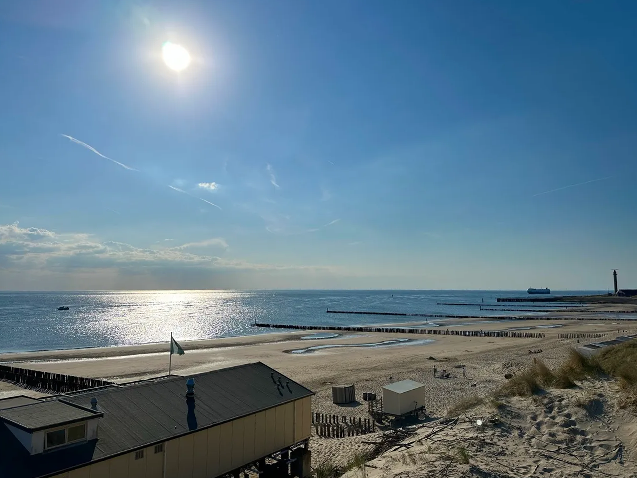 Sonniger Strand mit blauem Himmel, Wellen, Restaurant, Sand und Leuchtturm in der Ferne.