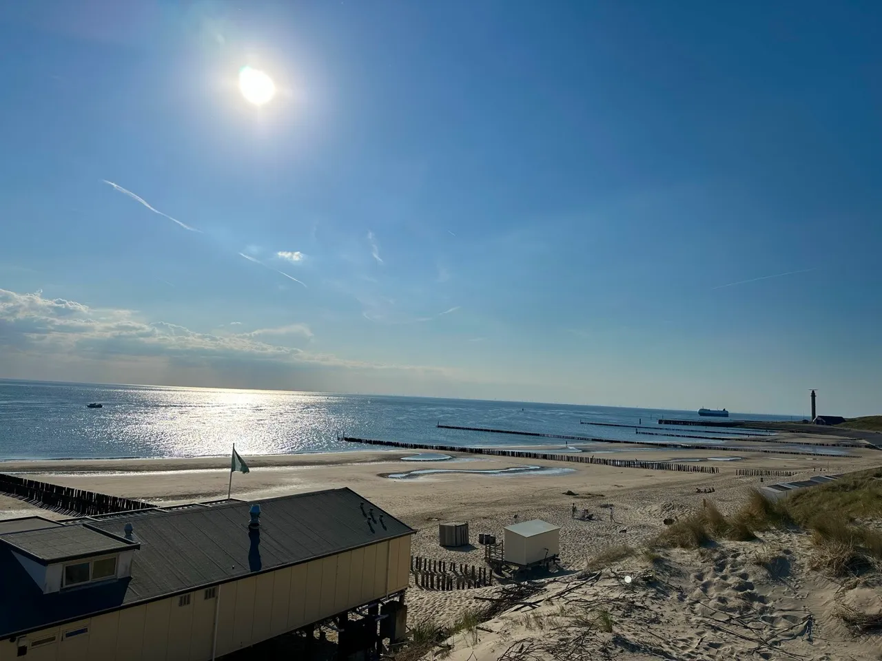 Sonniger Strand mit blauem Himmel, Wellen, Restaurant, Sand und Leuchtturm in der Ferne.