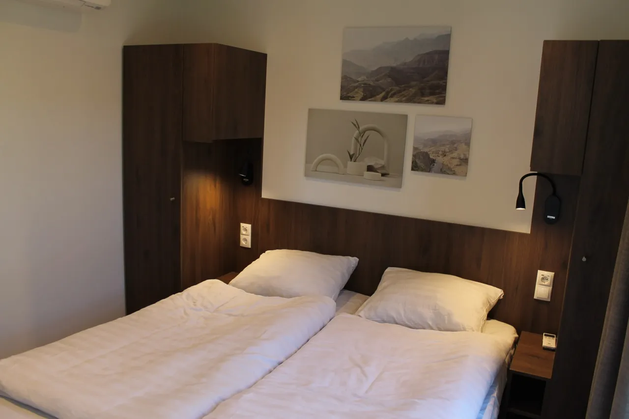 Doppelbett mit weißer Bettwäsche, Holzschränken, Leselampen, Wandbildern und Klimaanlage.
