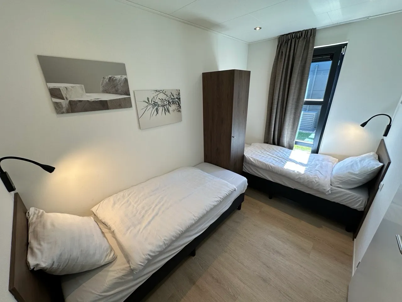 Helles Zimmer mit zwei Einzelbetten, Holzschrank, Leselampen, Bildern und Fenster mit Vorhängen.