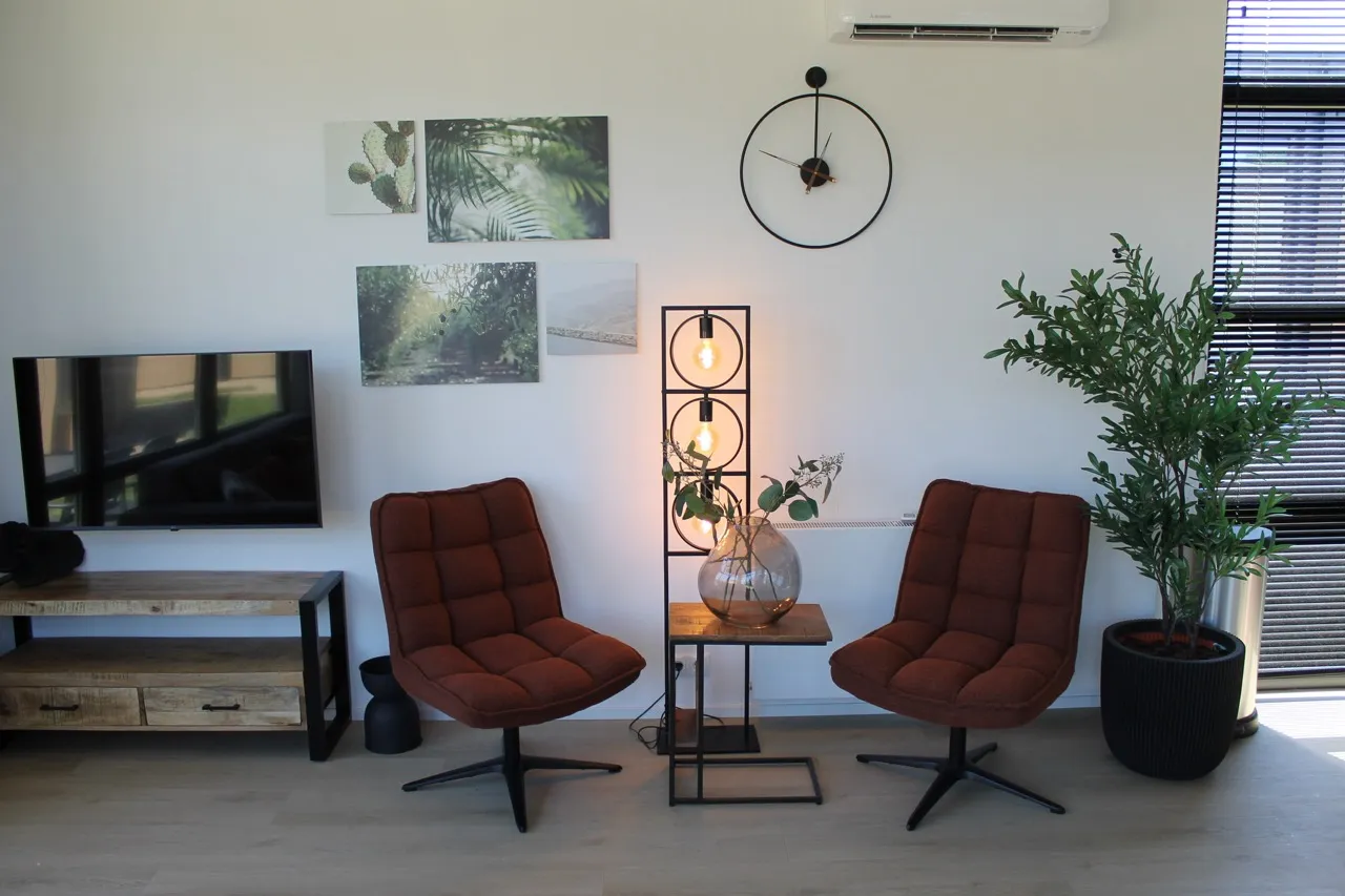 Wohnzimmer mit braunen Sesseln, Tisch, Lampe, TV,-Wand, Uhr, Pflanze, Fenster und Fenster mit Jalousien.