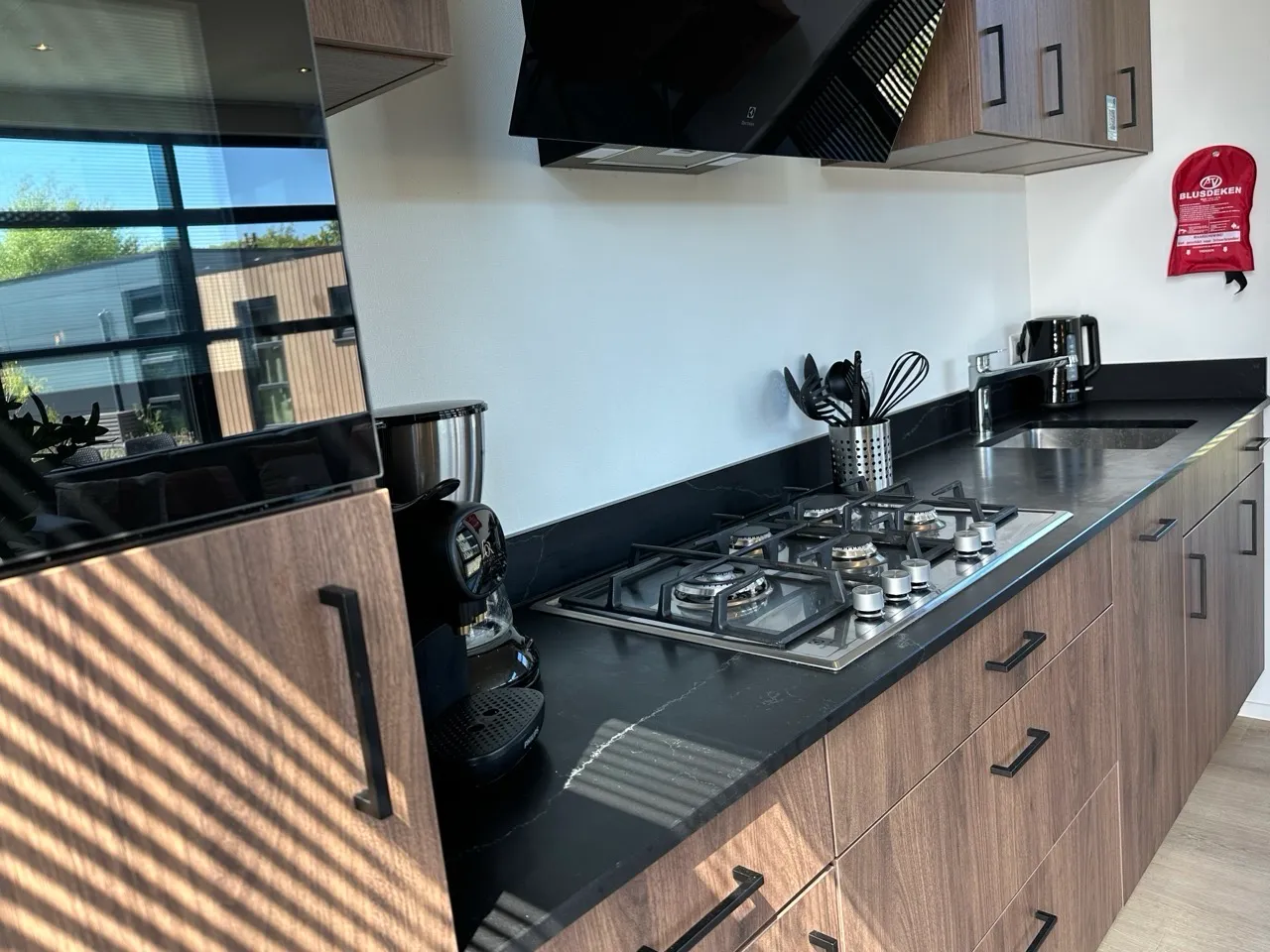 Küche mit Holzschränken, schwarzer Arbeitsplatte, Gasherd, Kaffeemaschine und Feuerdecke an der Wand.