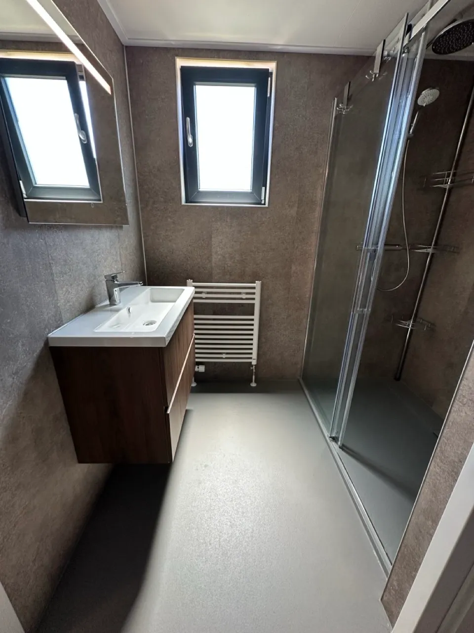 Modernes Bad mit Wandwaschbecken, Schrank, Glaswand, Regendusche, Heizkörper und Tageslicht.