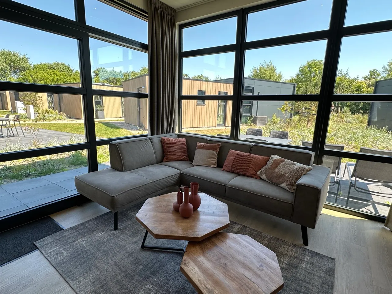 Modernes Wohnzimmer mit grauem L-Sofa, Couchtischen, hellen Kissen und Fenstern mit Blick ins Grüne.