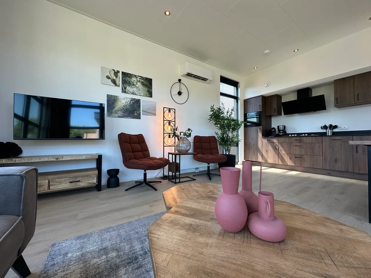 Wohnzimmer mit braunen Sesseln, Holztisch, rosa Vasen, Wand-TV, dunkler Küchenzeile und Bildern.
