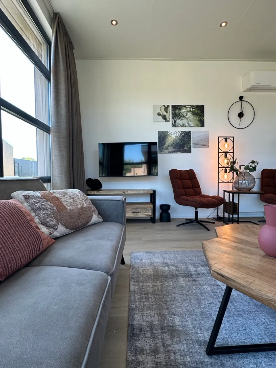 Helles Wohnzimmer mit grauer Couch, Holztisch, zwei Stühlen, Lampe, Wand-TV, Kunst und großen Fenstern.
