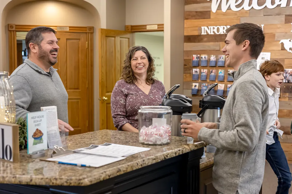 Three adults smile and chat at a welcome desk with coffee pots and a jar of mints.
