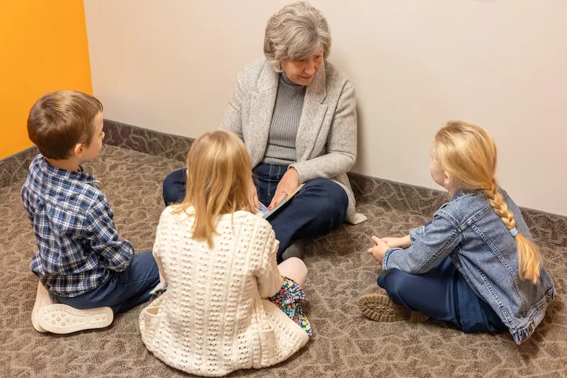 A woman sits on the floor reading a book to three kids gathered around her.
