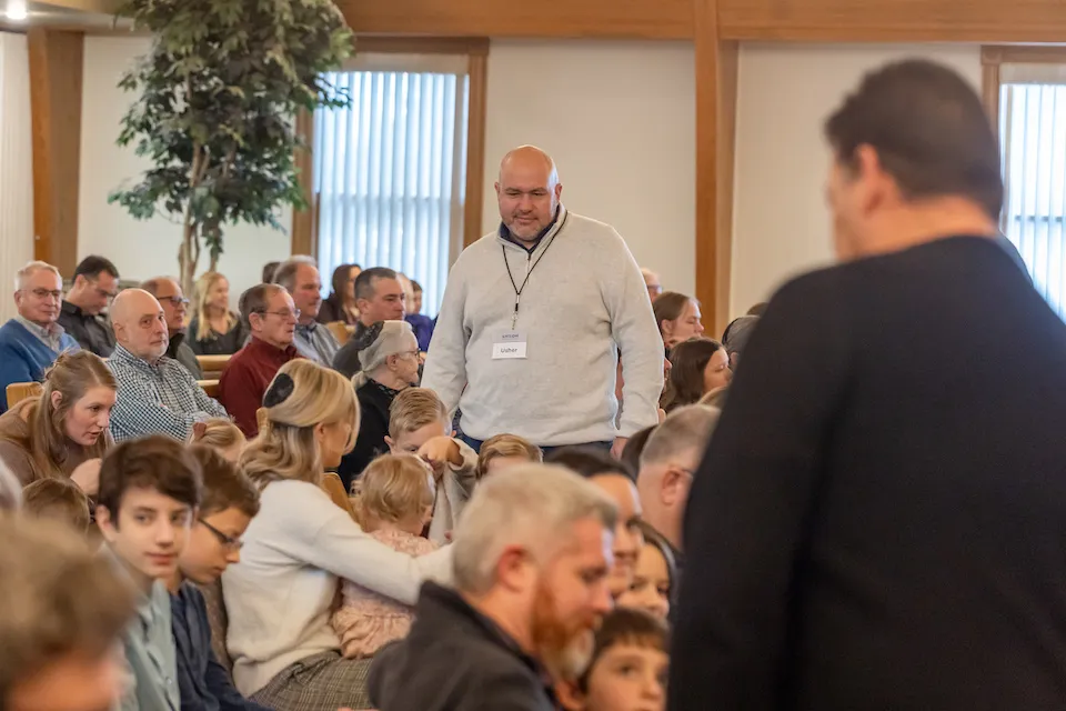 An usher wearing a sweater walks through a crowd of people seated in the sanctuary.