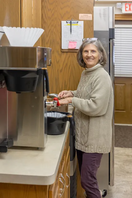 A smiling woman with gray hair in a cable-knit sweater prepares coffee in a cozy kitchen.