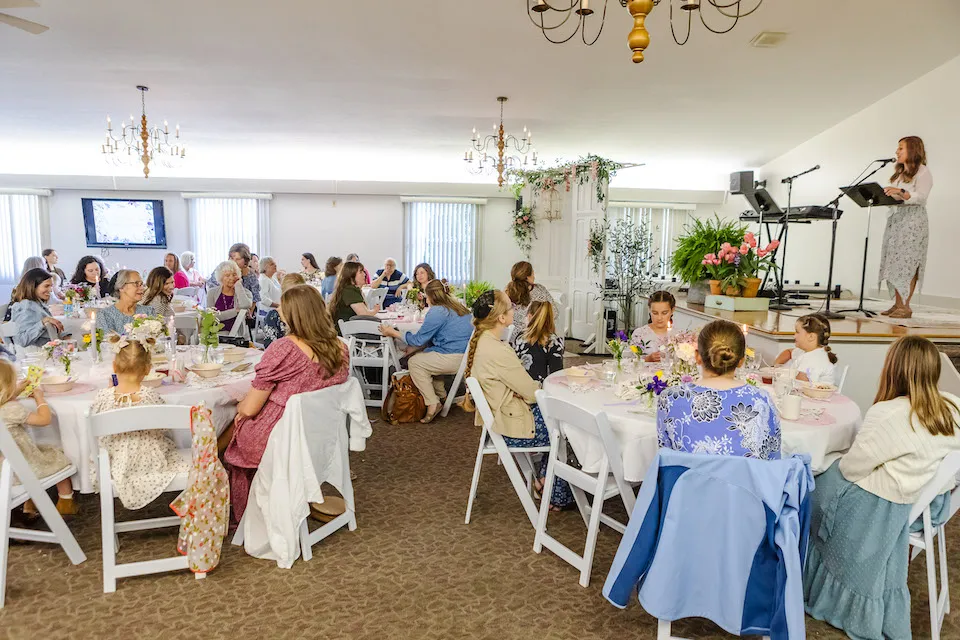 A gathering of women seated at tables with flowers, a speaker at a podium, and a screen in the background.