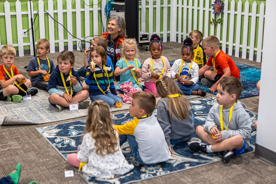 A group of young children sitting on rugs in a classroom, listening attentively with adult supervision.