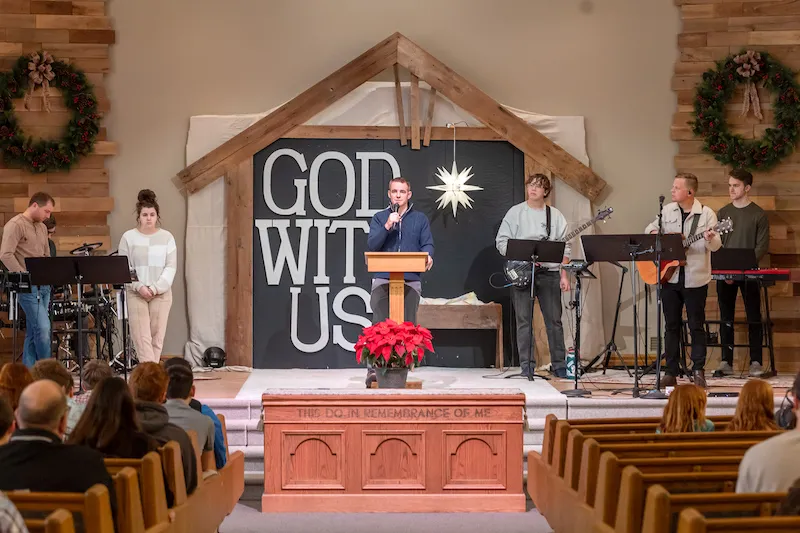 A worship service with a speaker at a podium, musicians playing instruments, and a backdrop that reads 'GOD WITH US', adorned with holiday decor.