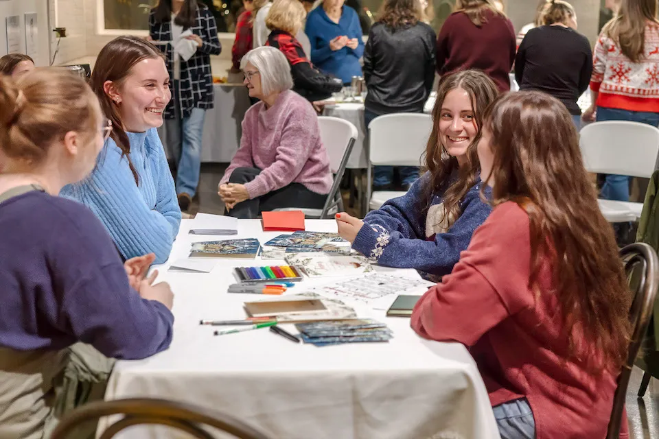 Four young women chatting at a table with art supplies and colorful drawings, enjoying a creative moment.