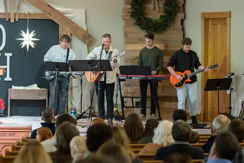 Musicians perform in front of an audience in a church setting, with instruments and decorations. 