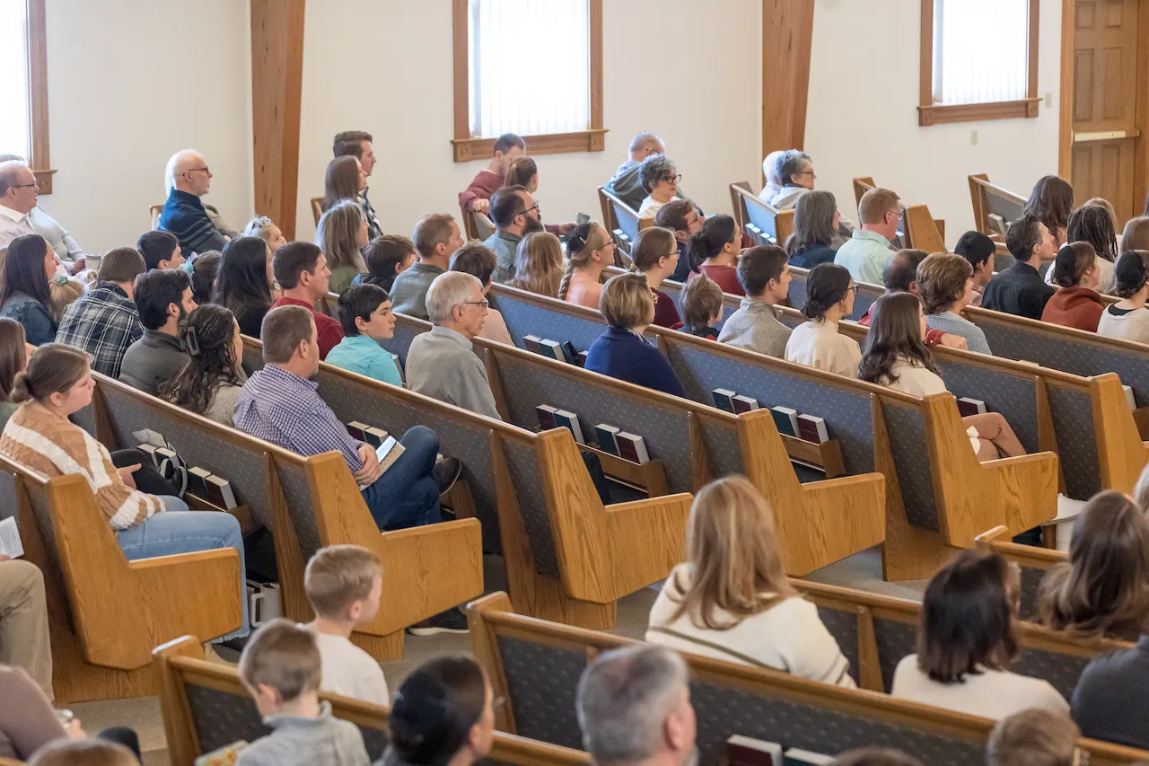 A crowd of people seated in church during a service, including adults and children, with wooden benches and windows. 