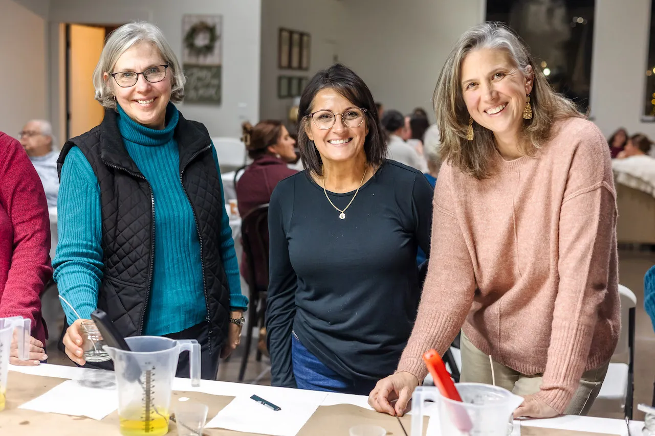 Three women smile while gathered around a table with measuring cups and bowls.