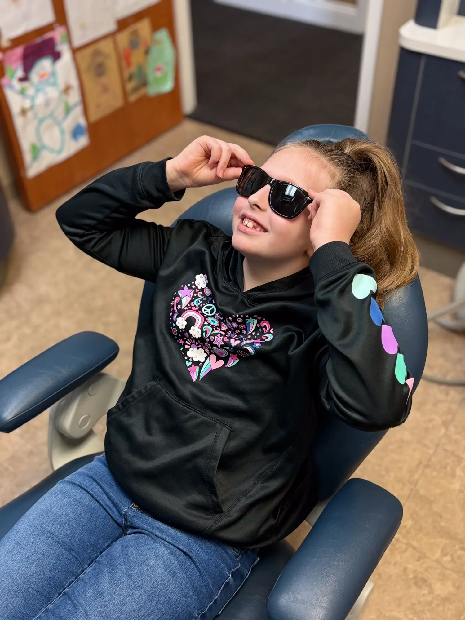 Smiling girl sitting in a dental chair wearing sunglasses and a black hoodie with colorful heart-shaped designs.