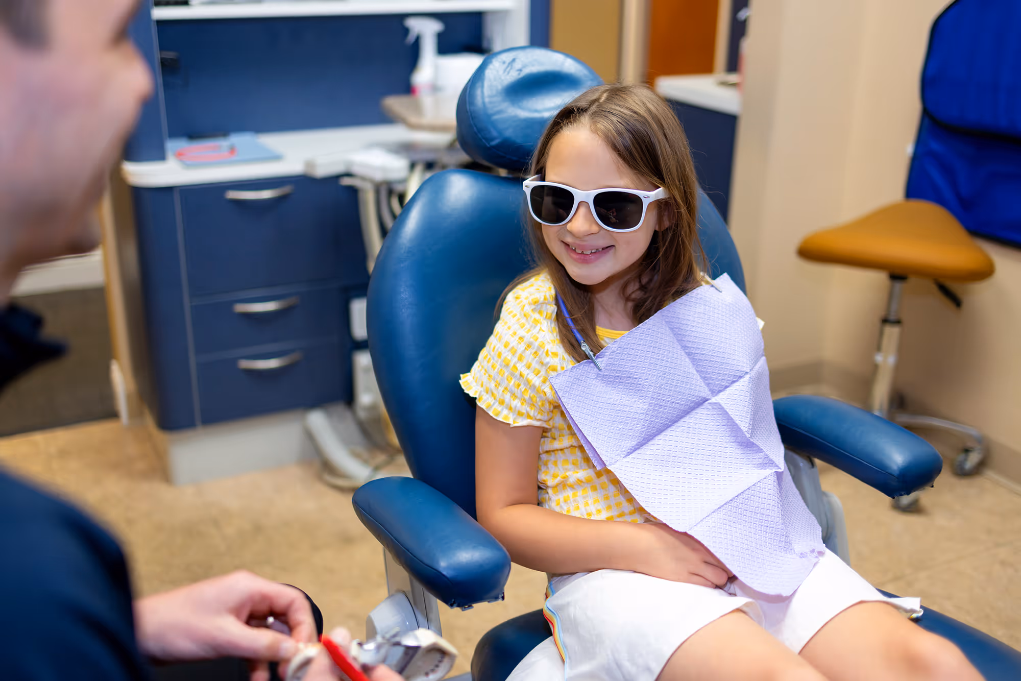 Smiling young girl wearing sunglasses sitting in a dental chair with a dental bib, facing a person holding a dental instrument.