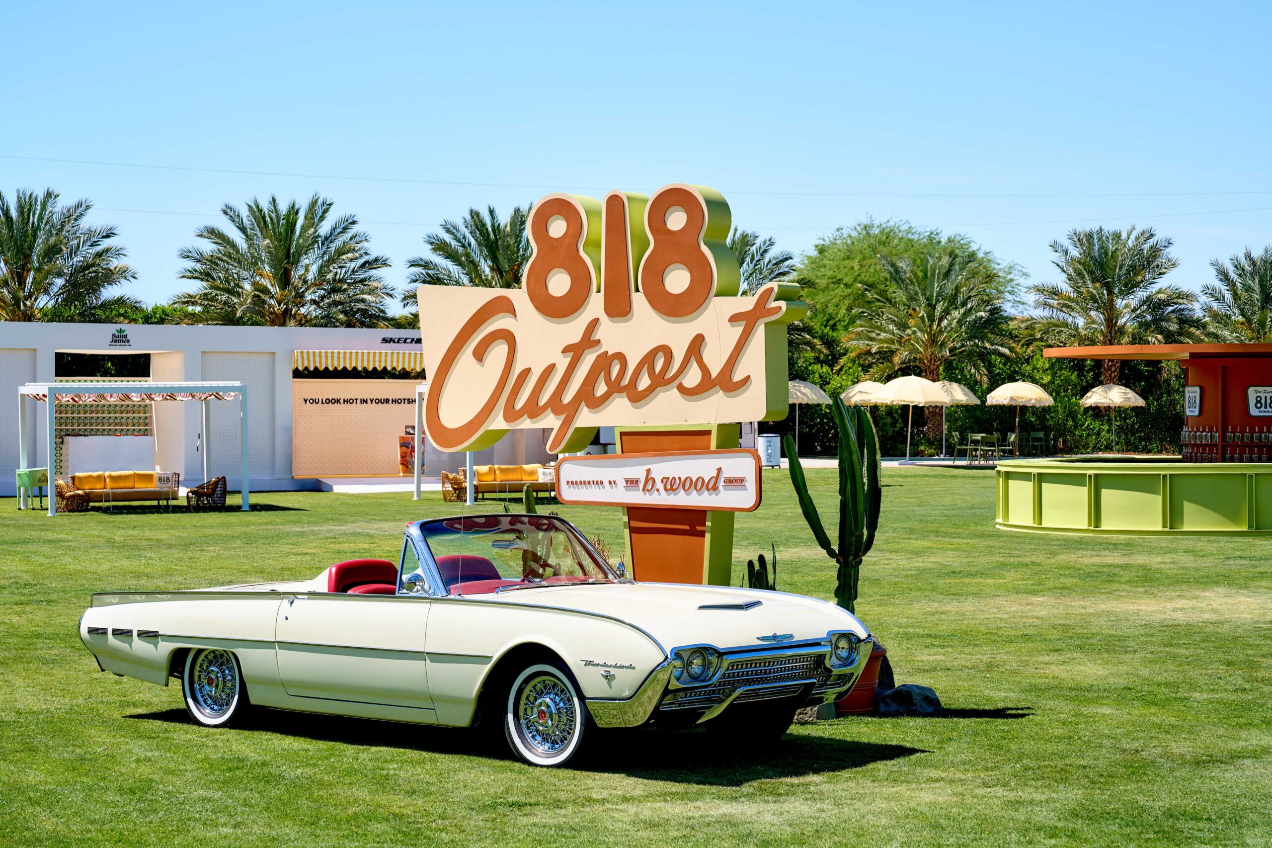 Classic white Ford Thunderbird convertible with red interior parked on grass near large 818 Outpost sign at a sunny outdoor event with palm trees.