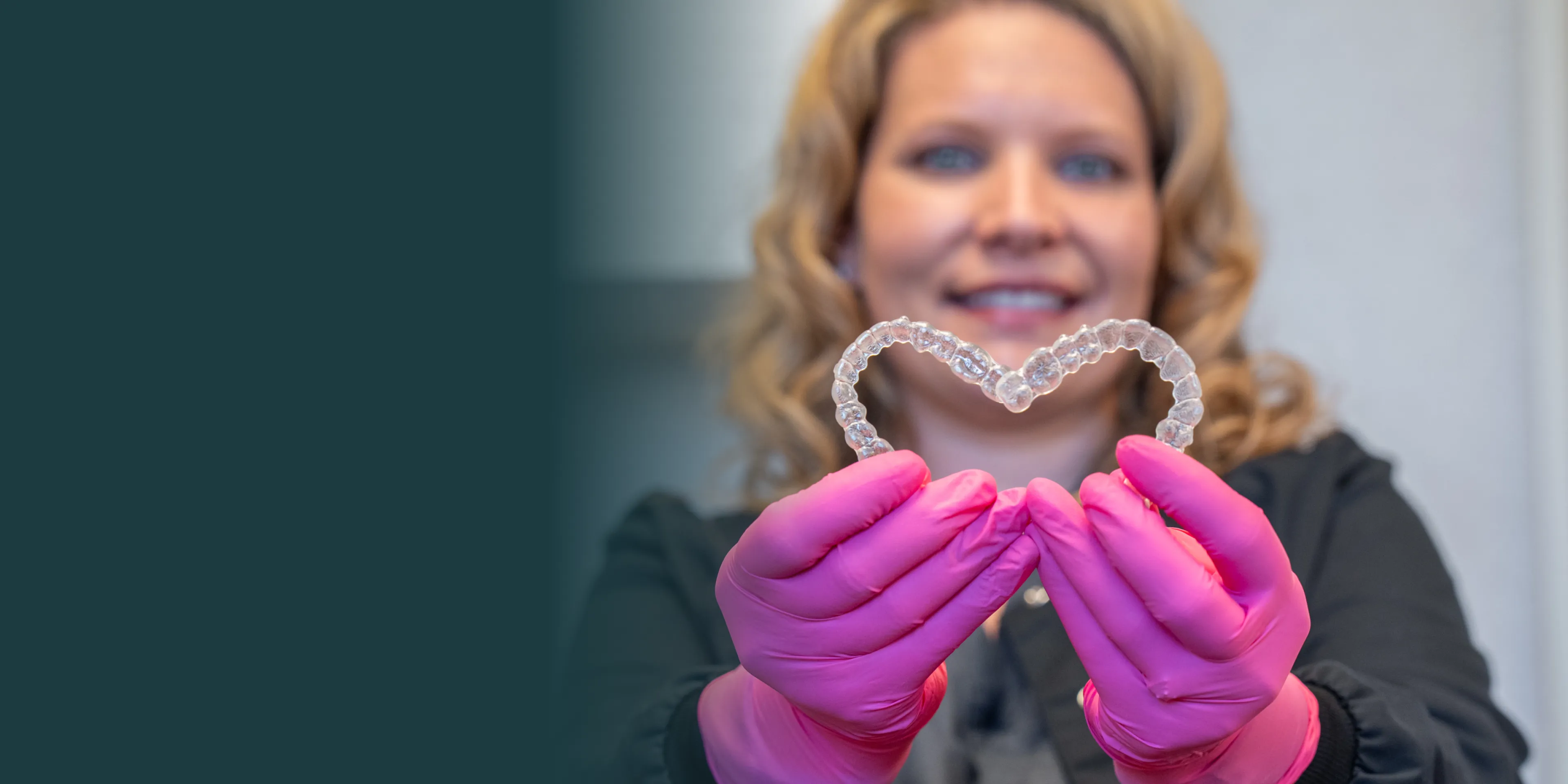 A person wearing pink gloves holds a heart-shaped dental aligner, smiling at the camera.