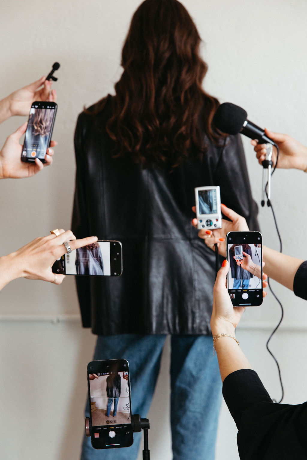 A girl with her back turned from the camera, with hands holding phones, cameras, and microphones trying to get her attention.