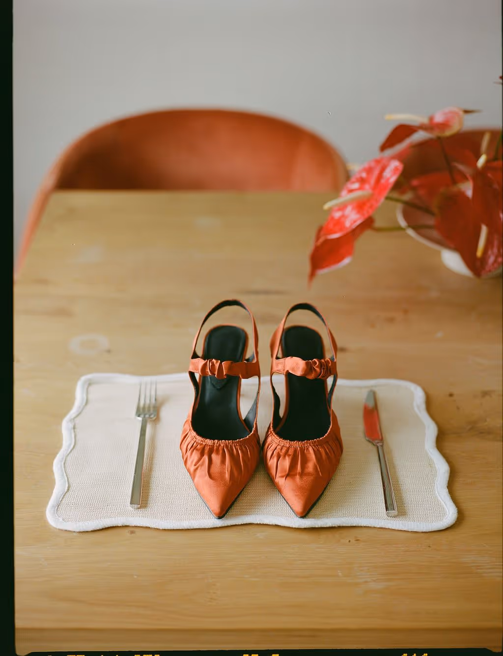 Pair of orange pointed-toe high heels placed side by side on a white placemat with a fork and knife on a wooden table.