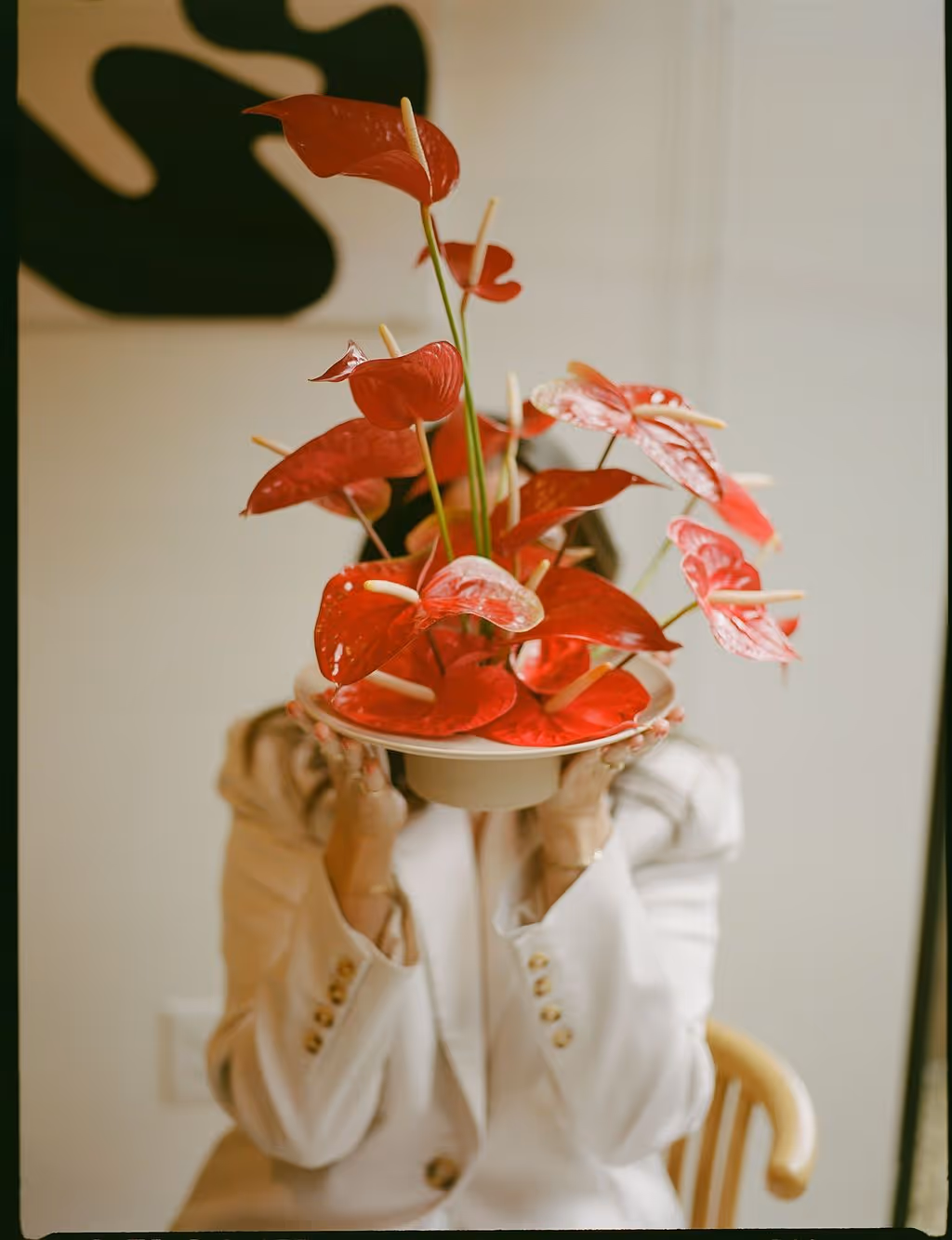 Person in a white blazer holding a plate with vibrant red anthurium flowers in front of their face.