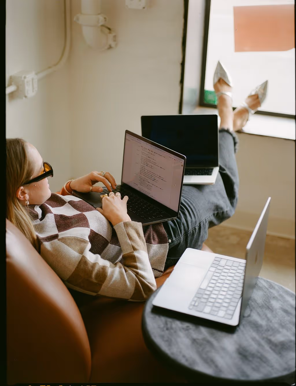 Woman wearing sunglasses lying on a couch working on a laptop with another laptop on a nearby table and a third laptop on her legs.