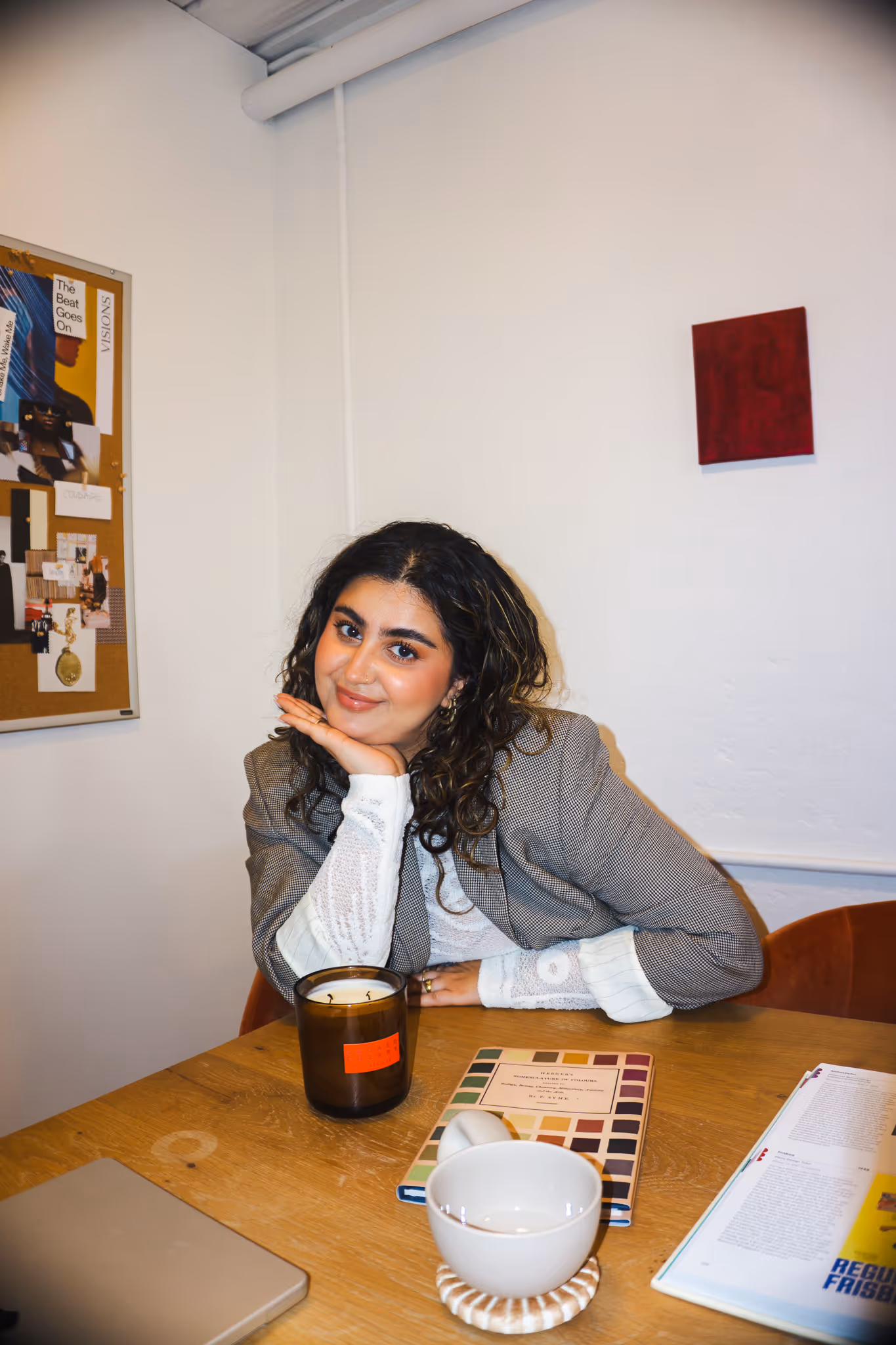 Woman with curly hair, wearing a gray blazer and white lace sleeves, sitting at a wooden table with a candle, a book, a white cup, and an open magazine.