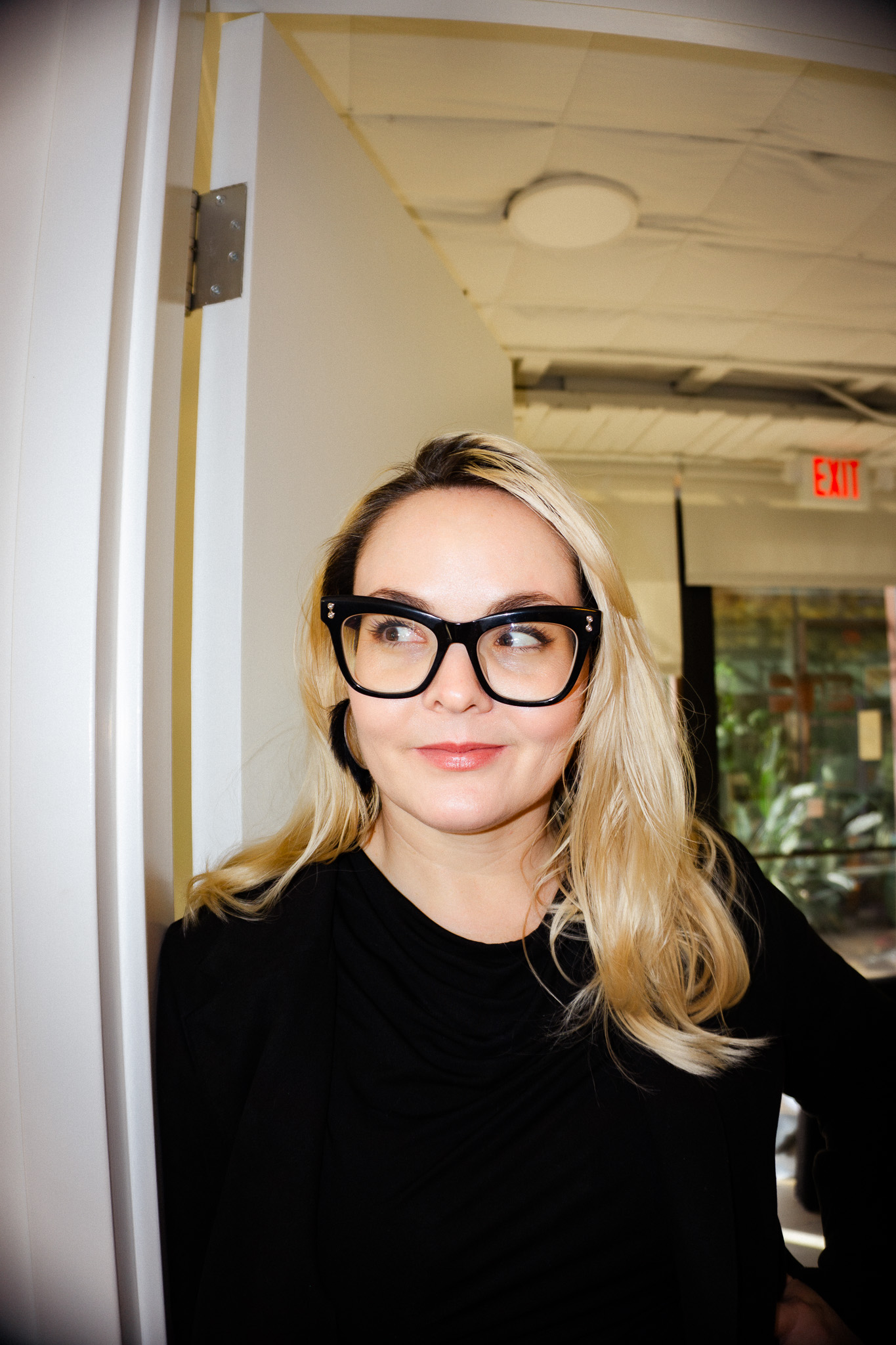 Blonde woman with black framed glasses wearing a black top, standing indoors near a white wall.