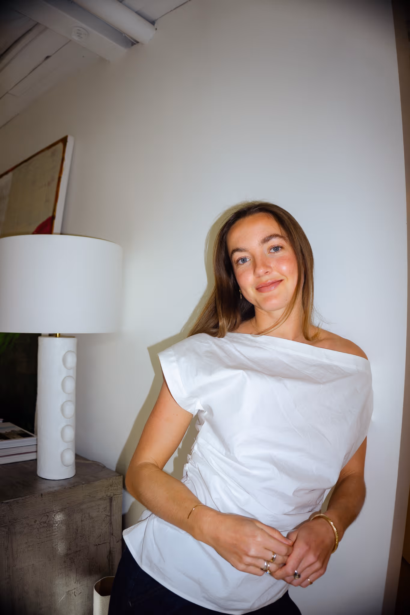 Smiling woman with long brown hair wearing a white off-the-shoulder blouse leaning against a wall near a modern white lamp on a wooden table.