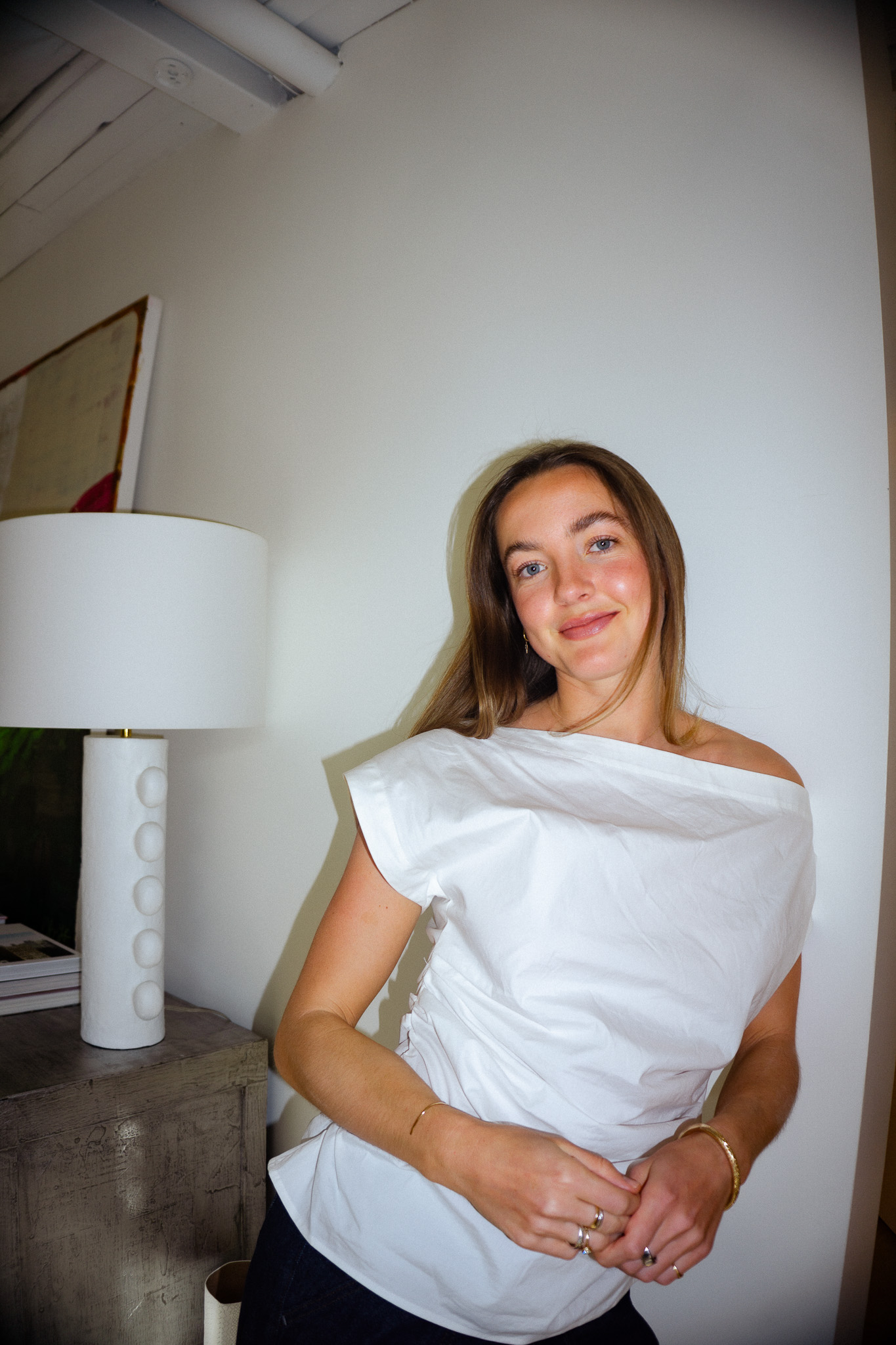 Smiling woman with long brown hair wearing a white off-the-shoulder blouse leaning against a wall near a modern white lamp on a wooden table.